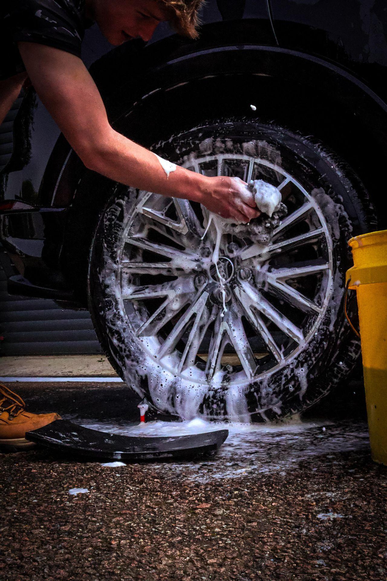 Person cleaning a car wheel with a foamy sponge beside a yellow bucket.