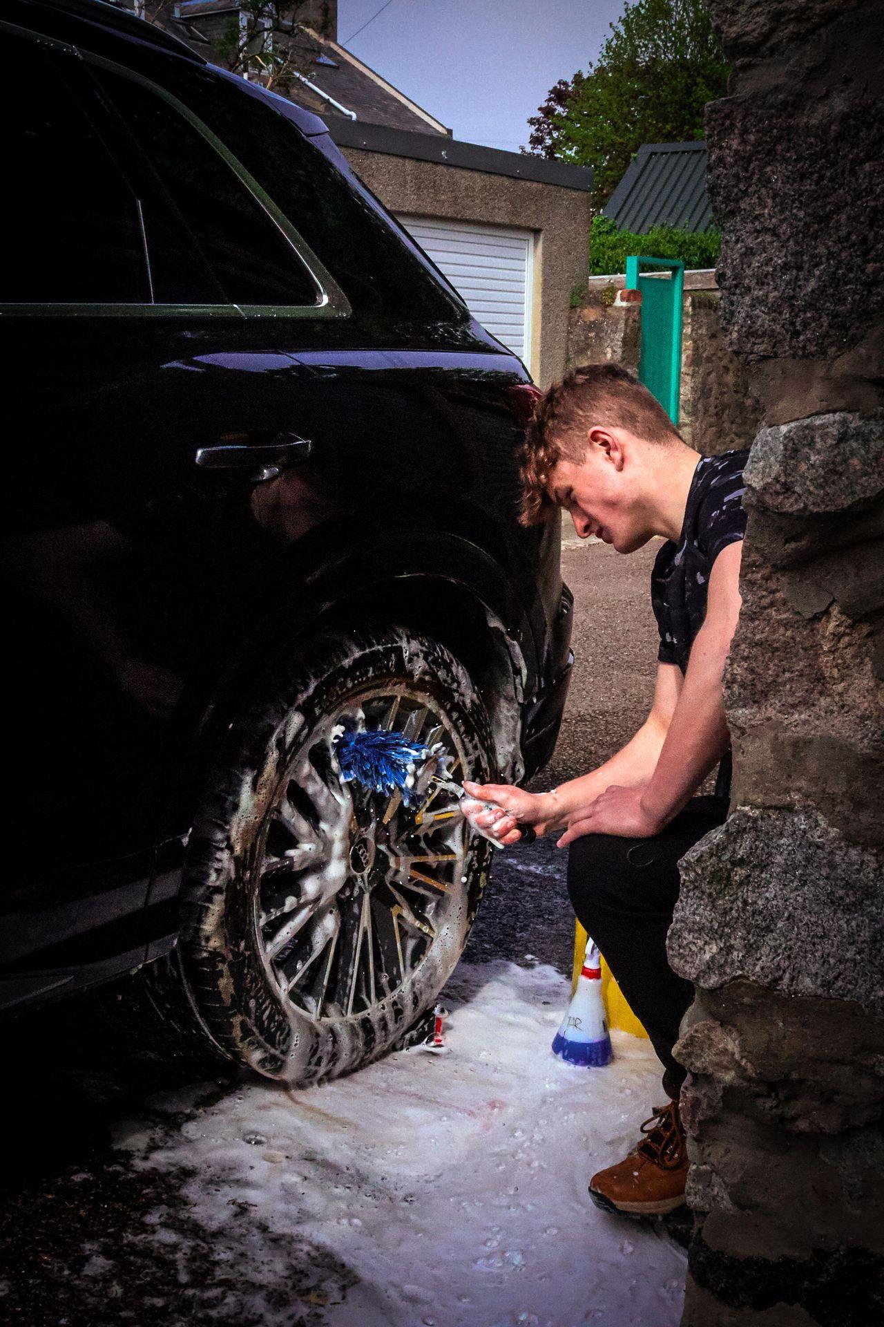 Person cleaning a car wheel with a blue brush, surrounded by soap and foam.