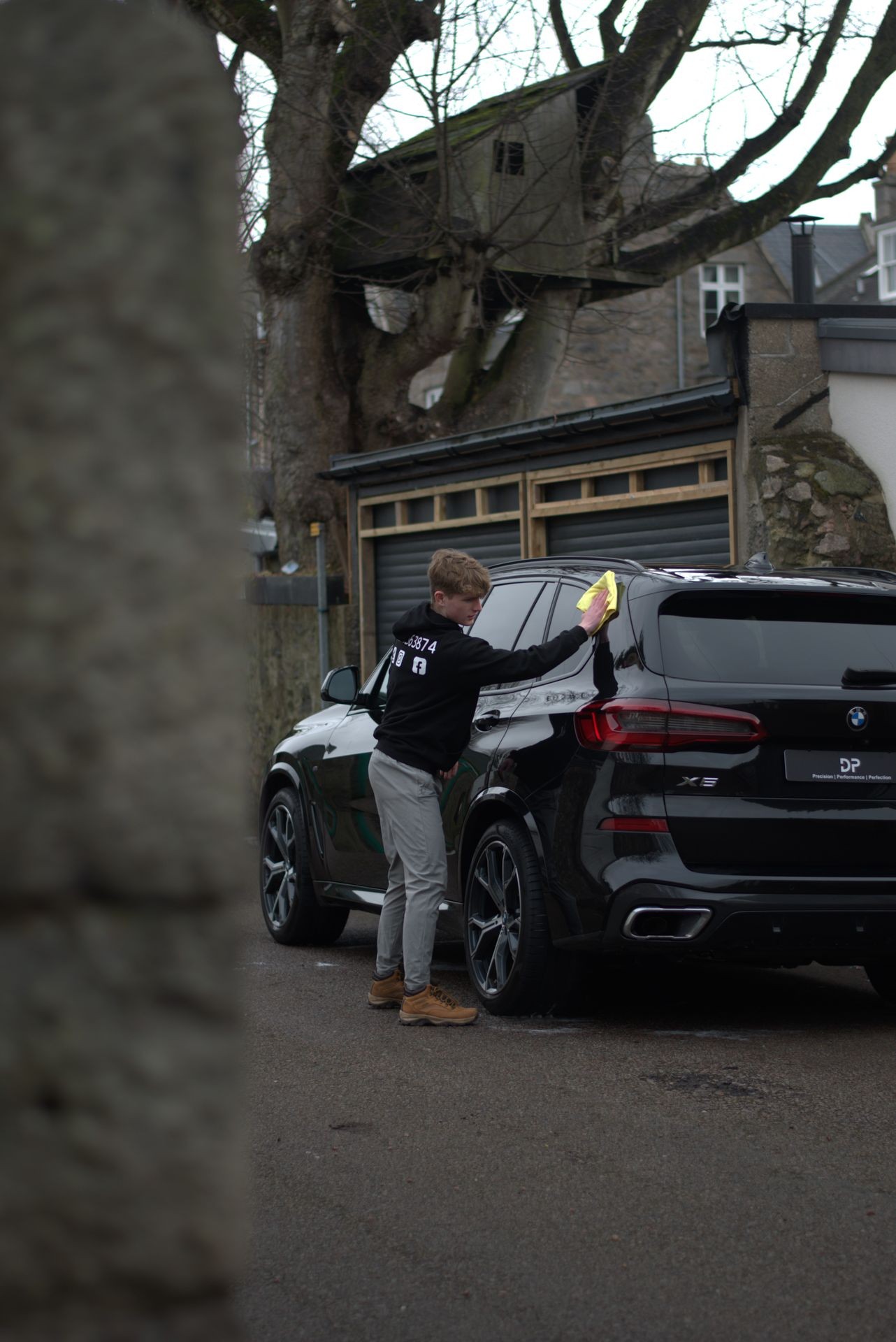 Person cleaning a black BMW X5 with a yellow cloth in front of a garage.