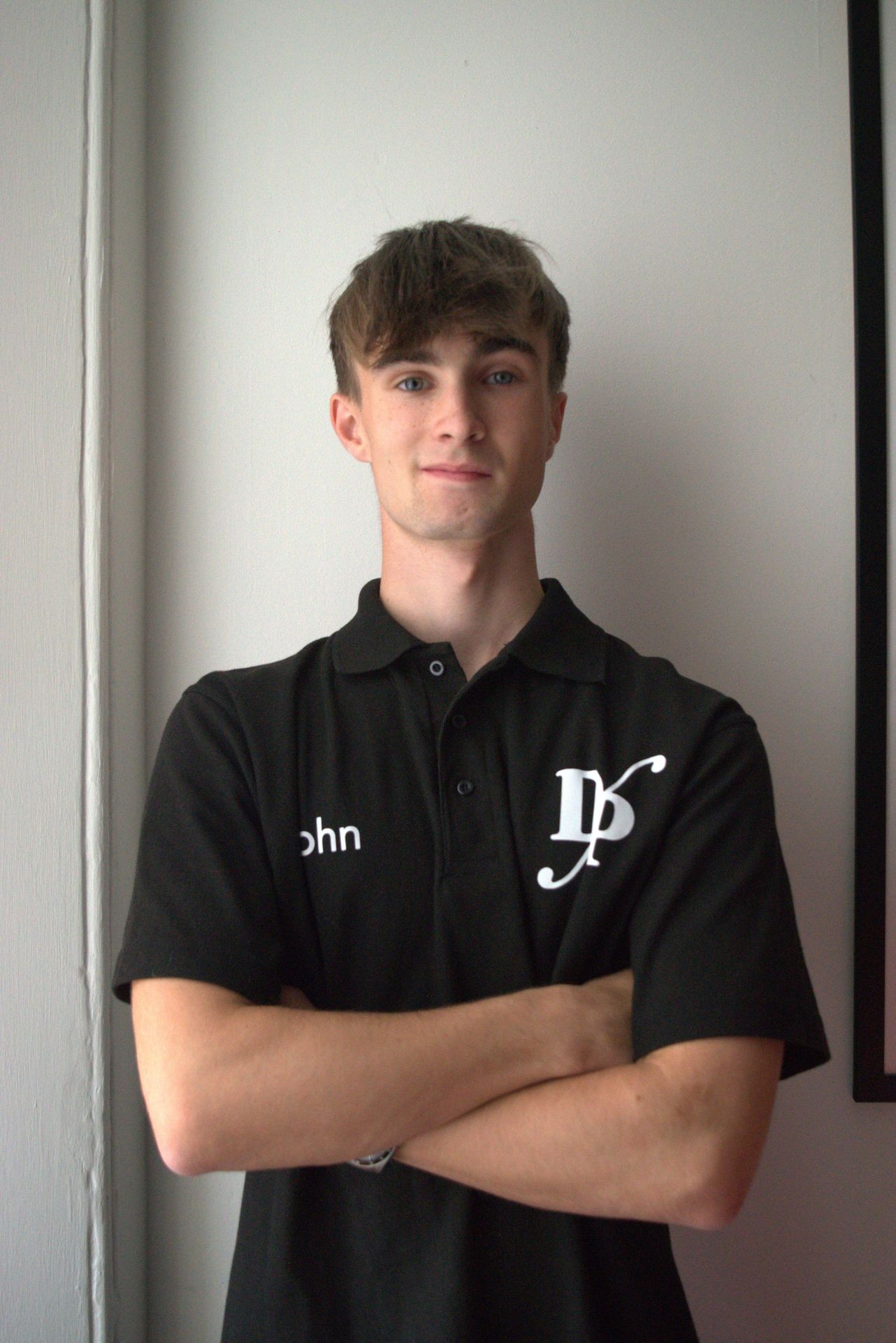 Young man in a black collared shirt with arms crossed, standing against a plain wall.