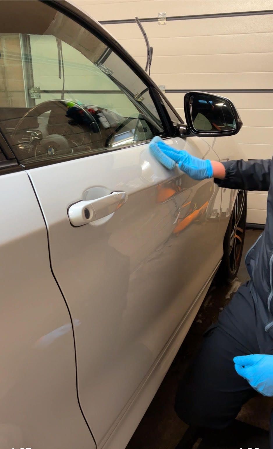 Person wearing blue gloves polishing a white car door in a garage.