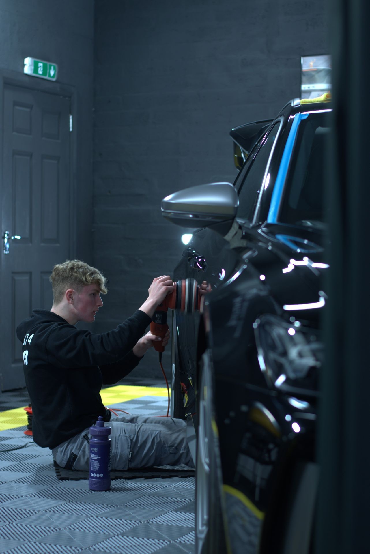 Person polishing the side of a black car with an electric polisher in a garage.