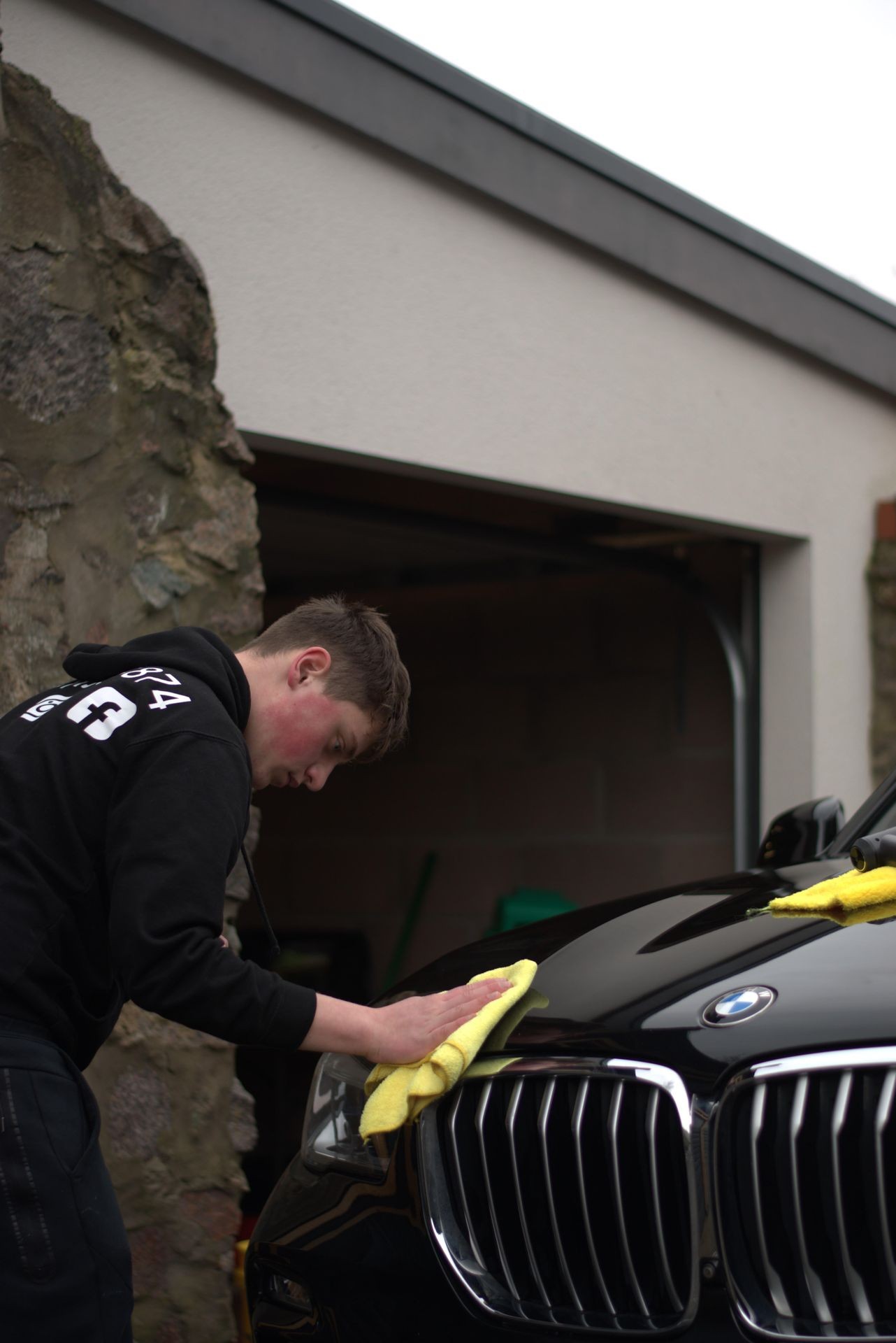 Person wearing a black hoodie polishing the front of a black car with a yellow cloth.