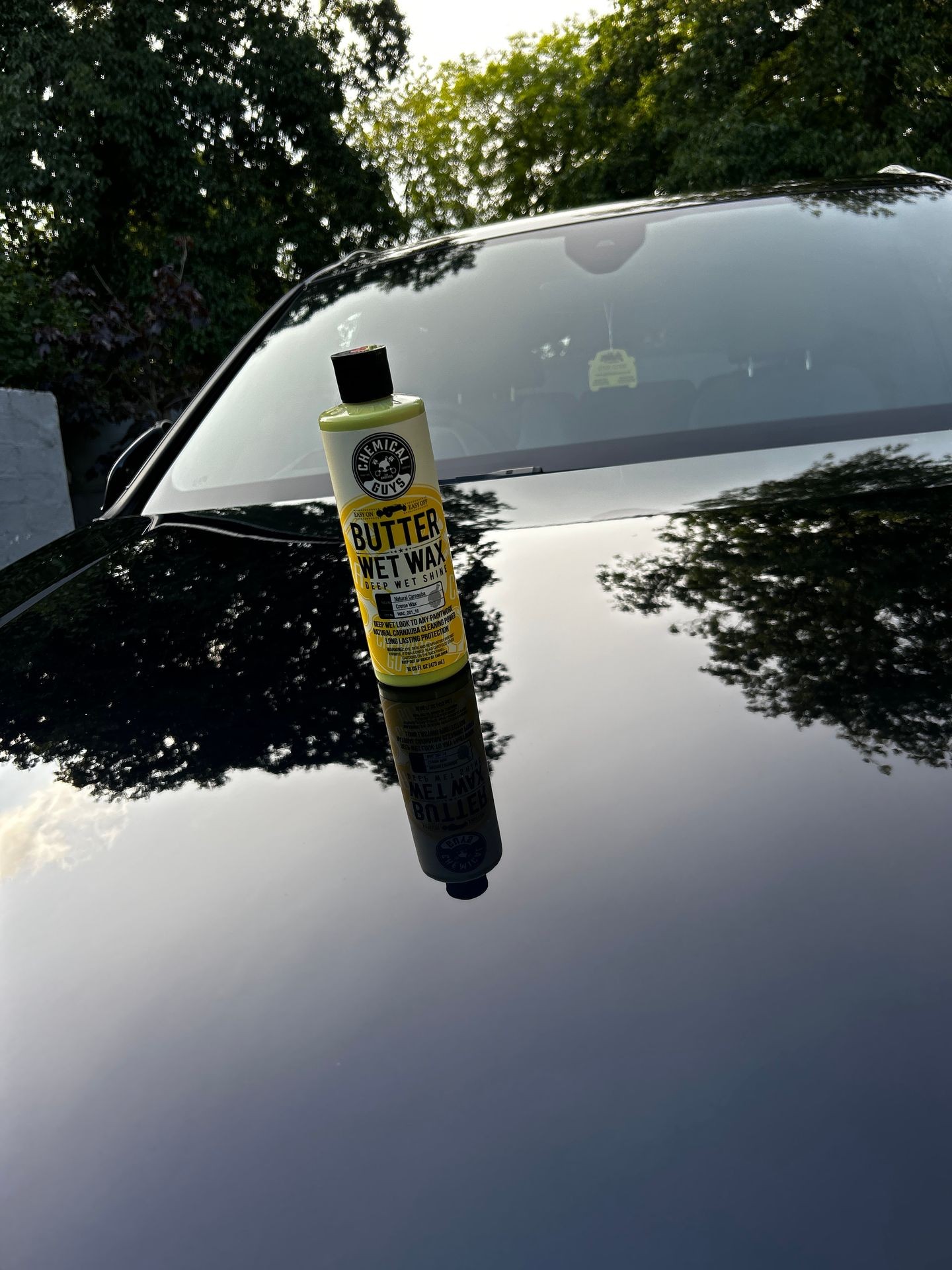 Bottle of car wax on a highly reflective black car hood with trees in the background.